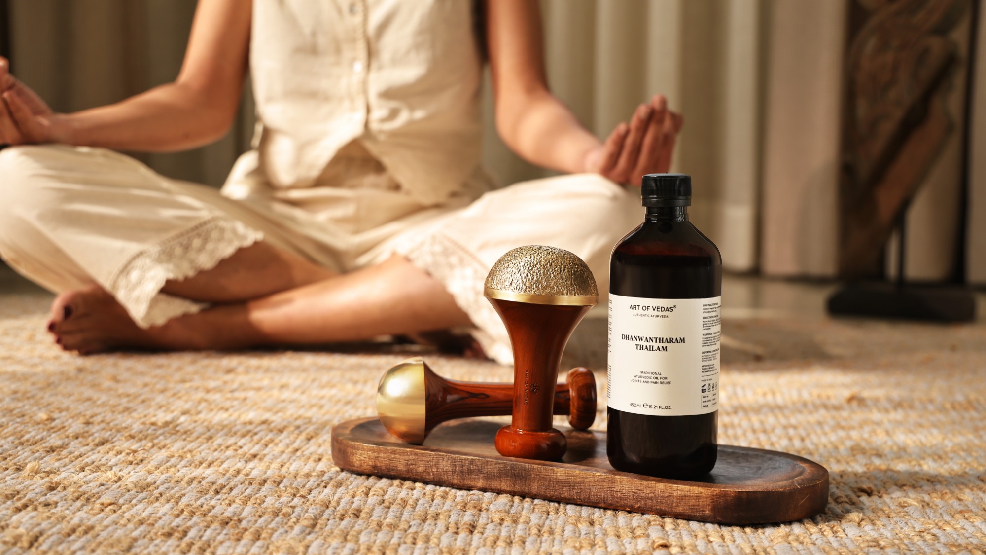 Twee Kansa wands van Art of Vedas en een fles Dhanwantharam Thailam op een houten dienblad, daarachter een vrouw in meditatie